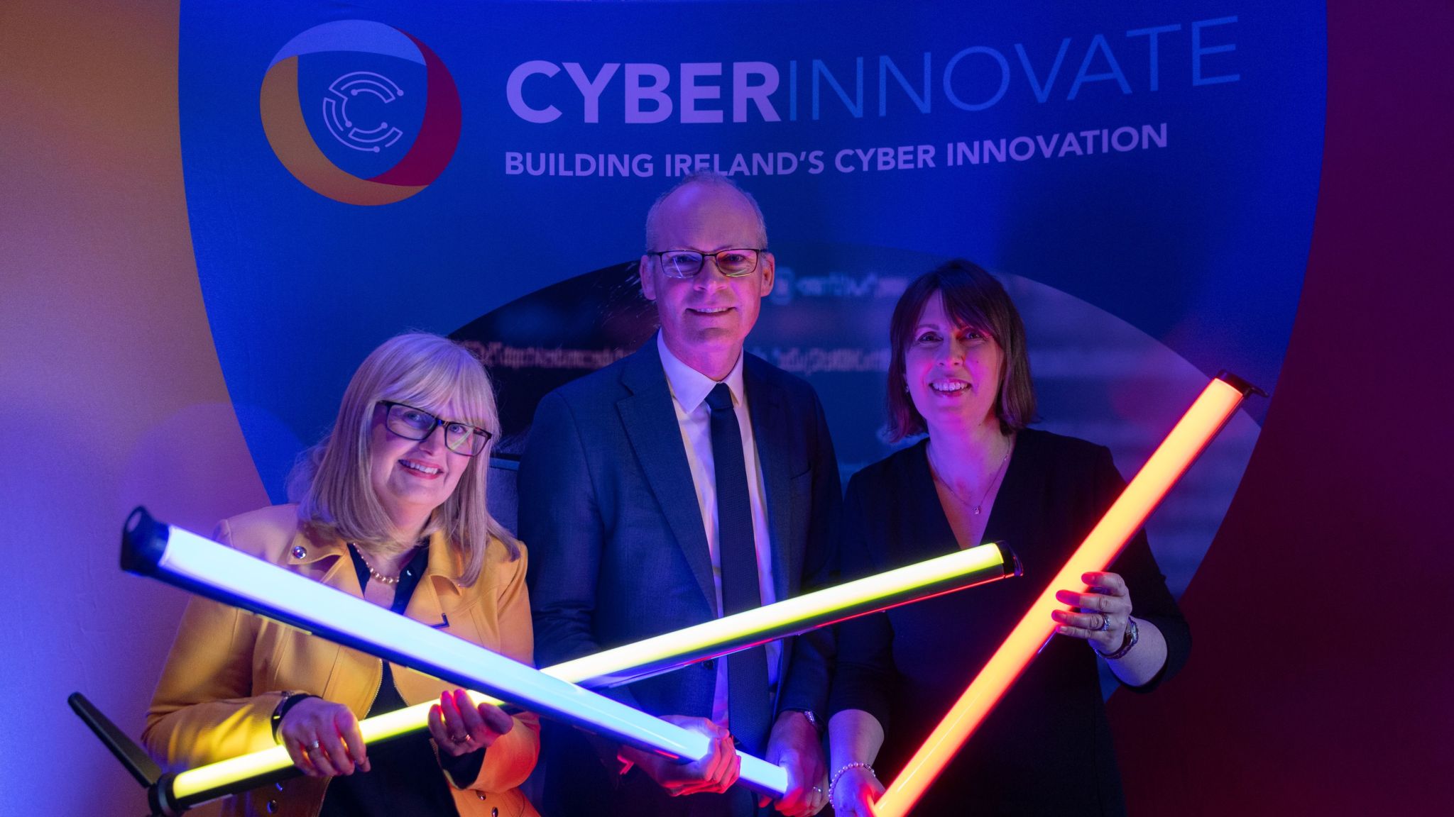 Donna OShea, Josette O'Mullane and Simon Coveney at the Cyber Innovate Launch holding up long LED lights in front of the Cyber Innovate banner in Cork jail.